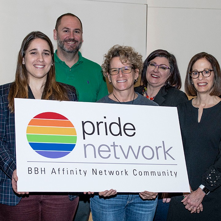 Five BBH'ers smiling at the camera holding a Pride Network BBH Affinity Network Community white sign with a rainbow circle on it