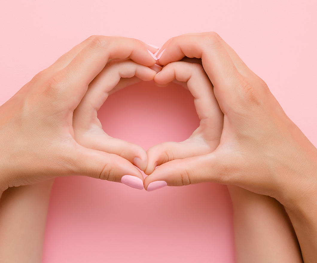 Adult hands forming a heart around a child's hand forming a heart on a pink background