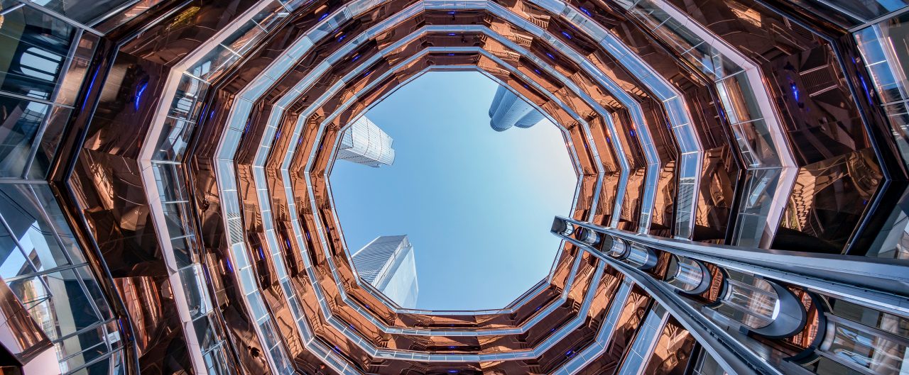 Hudson Yards wavy, modern structure taken from down below with a blue sky seen from the top and skyscrapers looming above