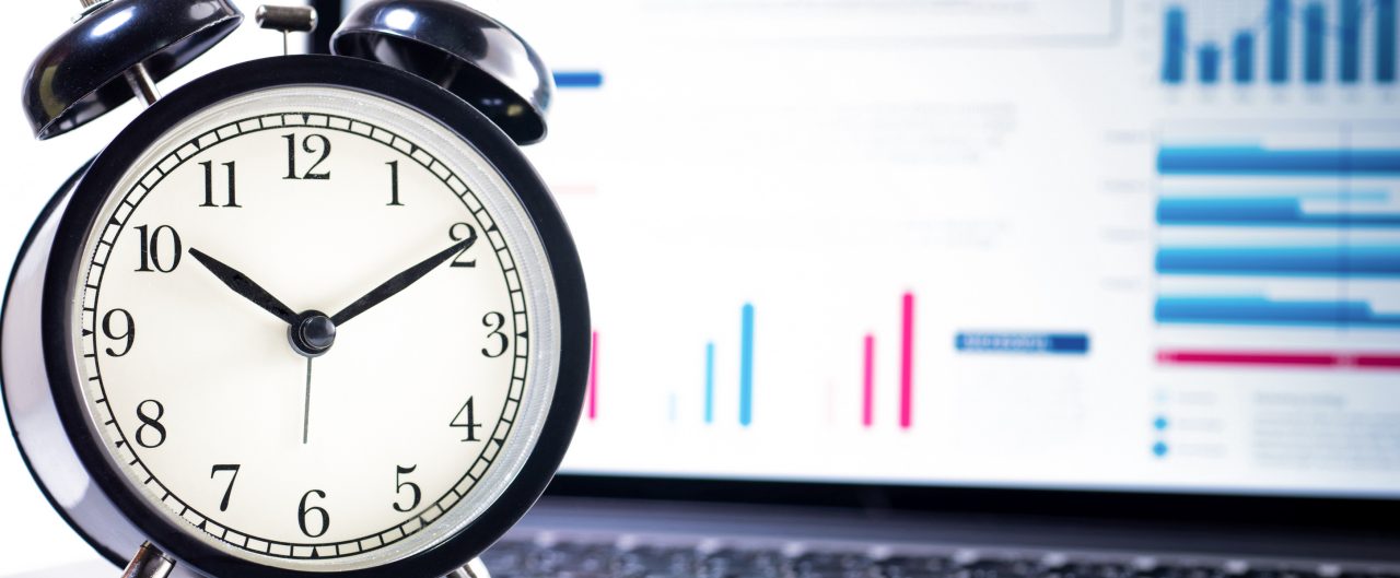 Black and white alarm clock standing next to an open laptop which has financial charts and graphics on the screen