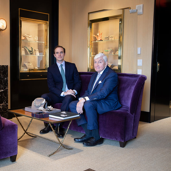 Two men sitting on a purple couch with glass jewelry display cases and marble fireplace 