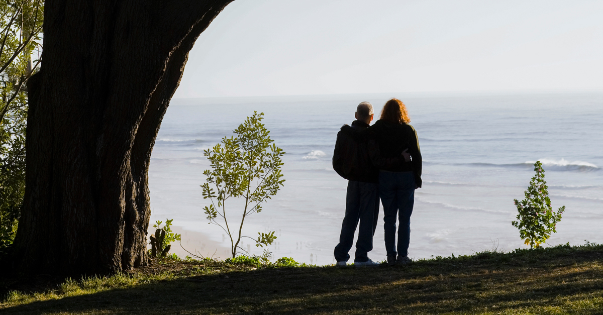 Couple Standing Under Tree With Backs To Camera Overlooking Ocean