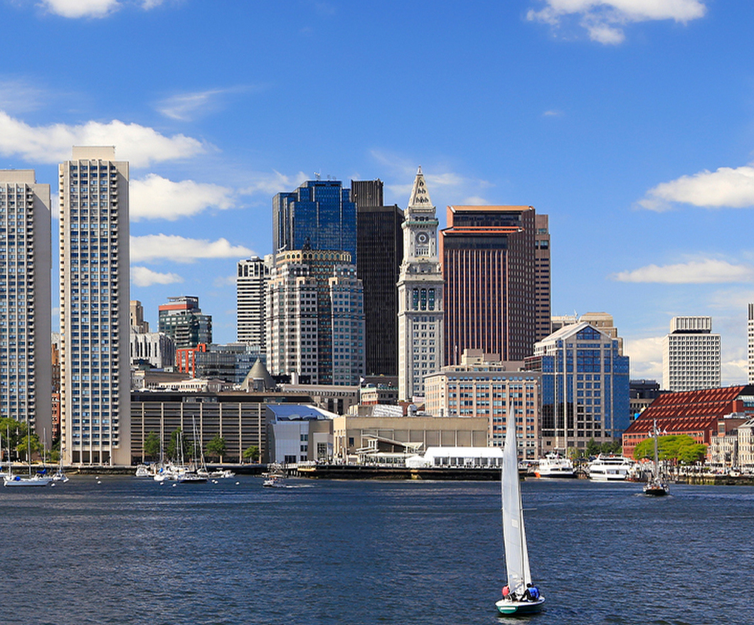 Boston city skyline seen behind a body of water with a sailboat in the center of the image