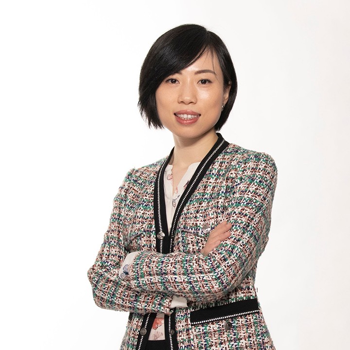 Smiling business woman with short black hair wearing a black, green and pink tweed blazer with arms crossed over her chest on a white backdrop