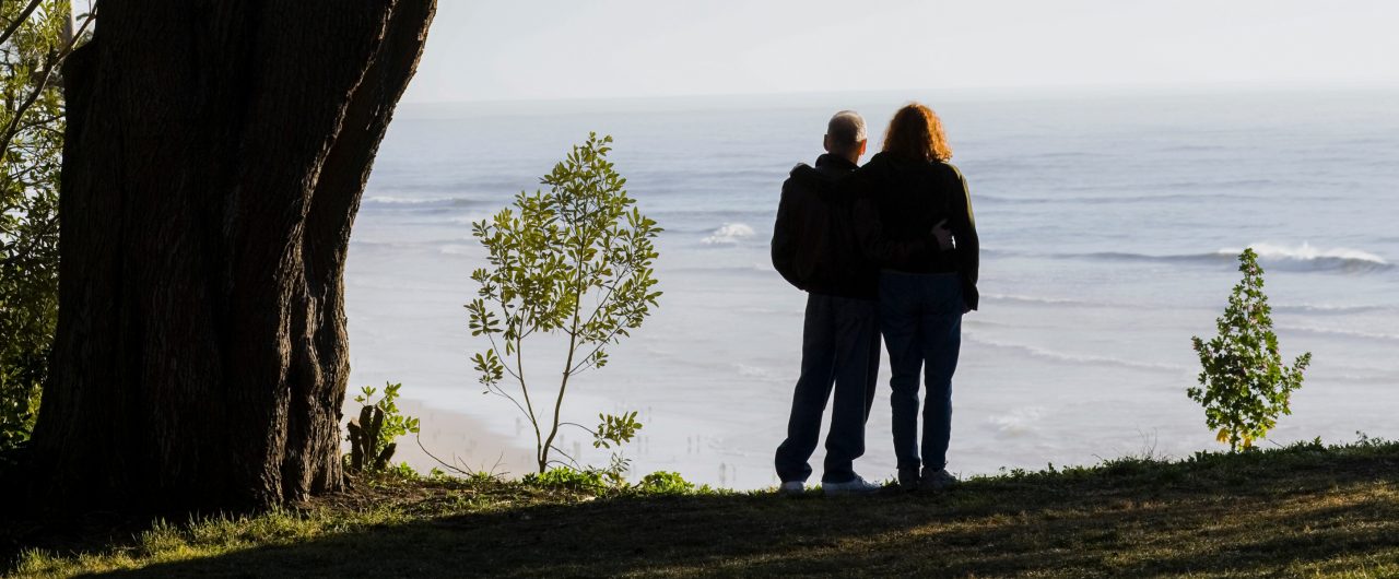 Couple Standing Under Tree With Backs To Camera Overlooking Ocean