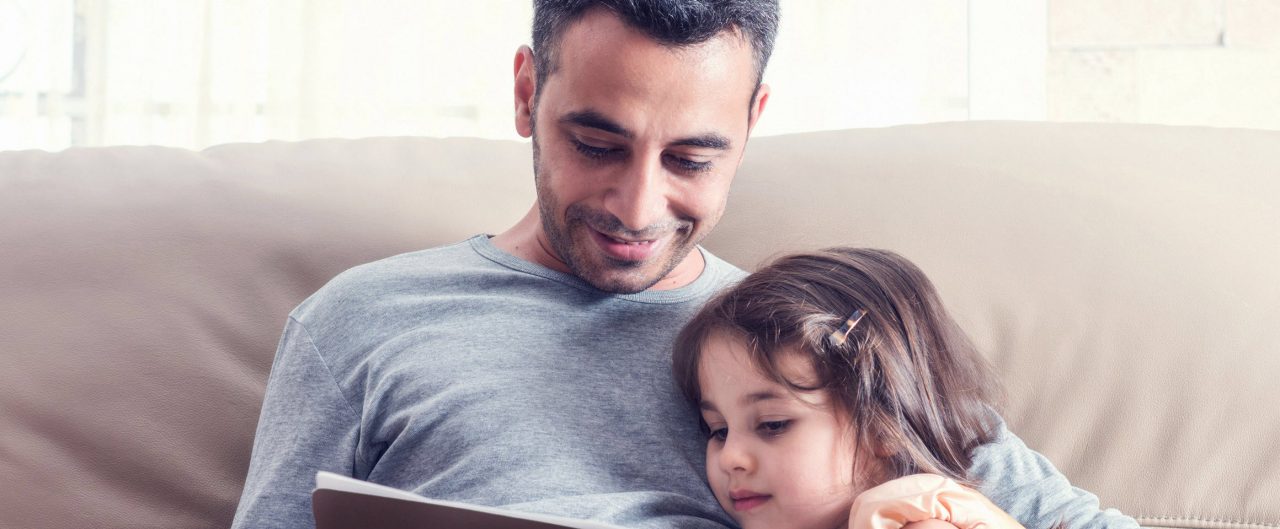 Little girl and father sitting together on a couch reading a book