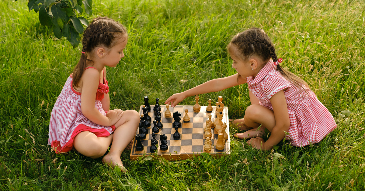 Two girls are playing chess in the garden on the grass. An old wooden chessboard with pieces. 