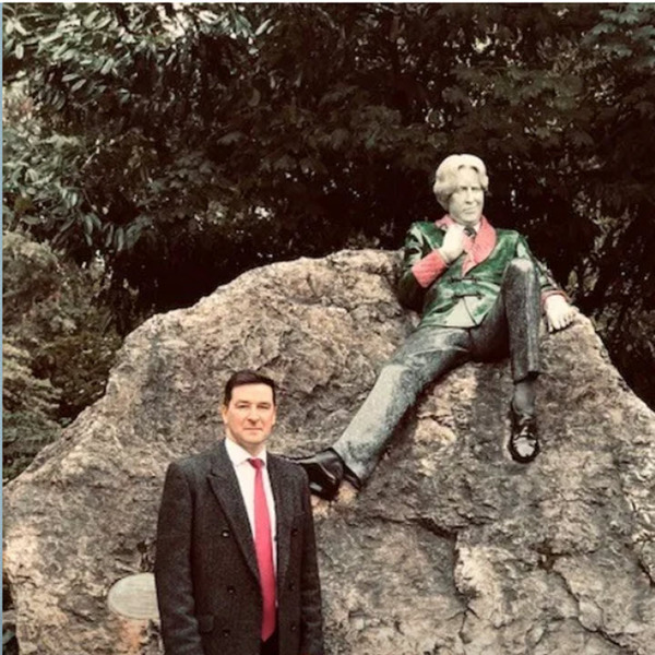 Man in suit standing next to statue of a man sitting on a boulder