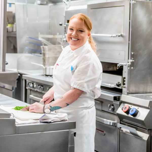 A female chef smiles and she chops greens up with a knife