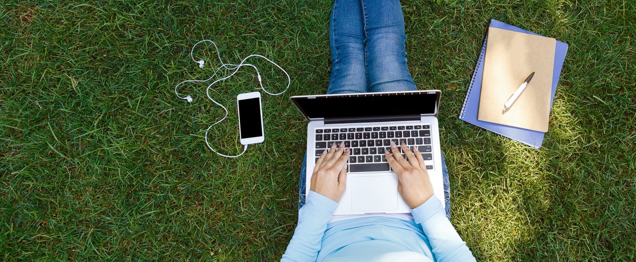 Top view of girl sitting in park on the green grass with laptop, notebook and smartphone with earphones,