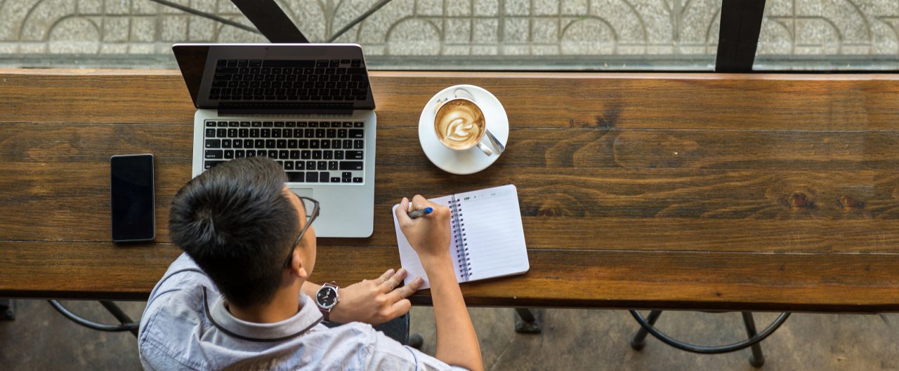 Young Asian worker taking notes beside laptop on wooden table; Remote work concept