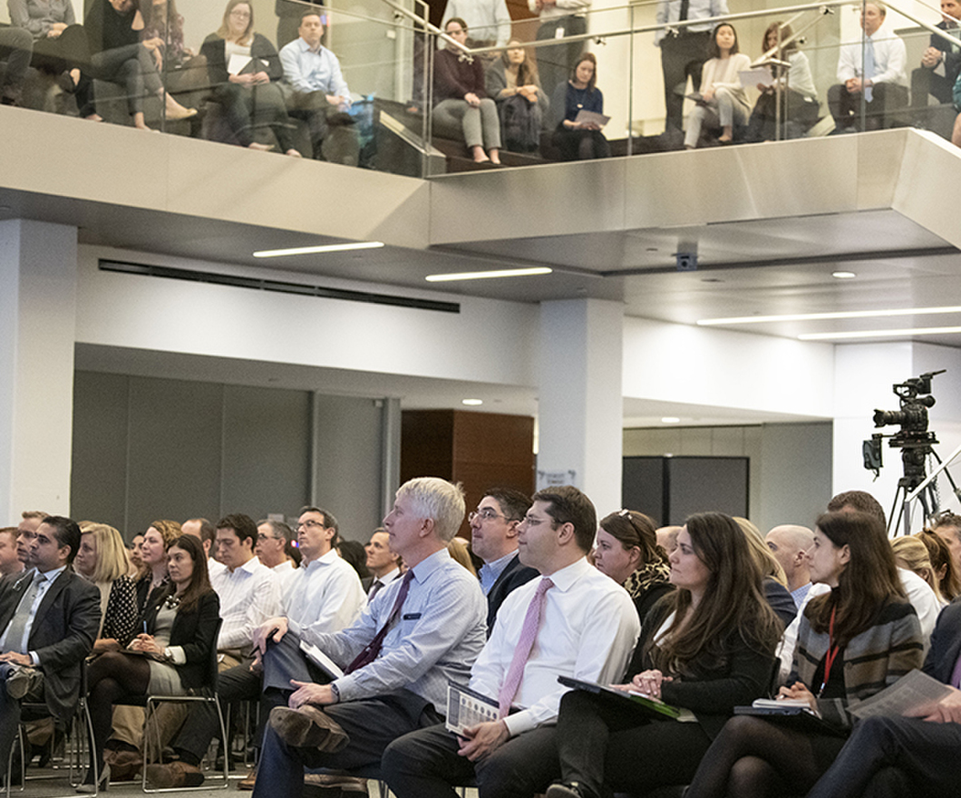 Audience of BBH'ers seen sitting in chairs on a first floor and standing on a second floor behind a glass wall