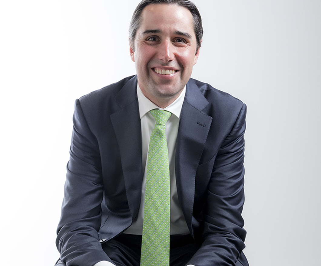 A smiling business man in a grey suit and green tie sitting with hands clasped on his thighs on a white background