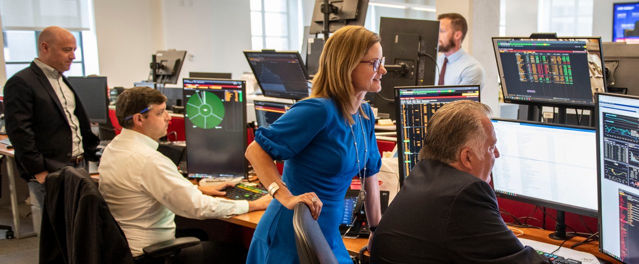 2 Tech BBH'ers sitting at their computers with 4 screens on the wall above them