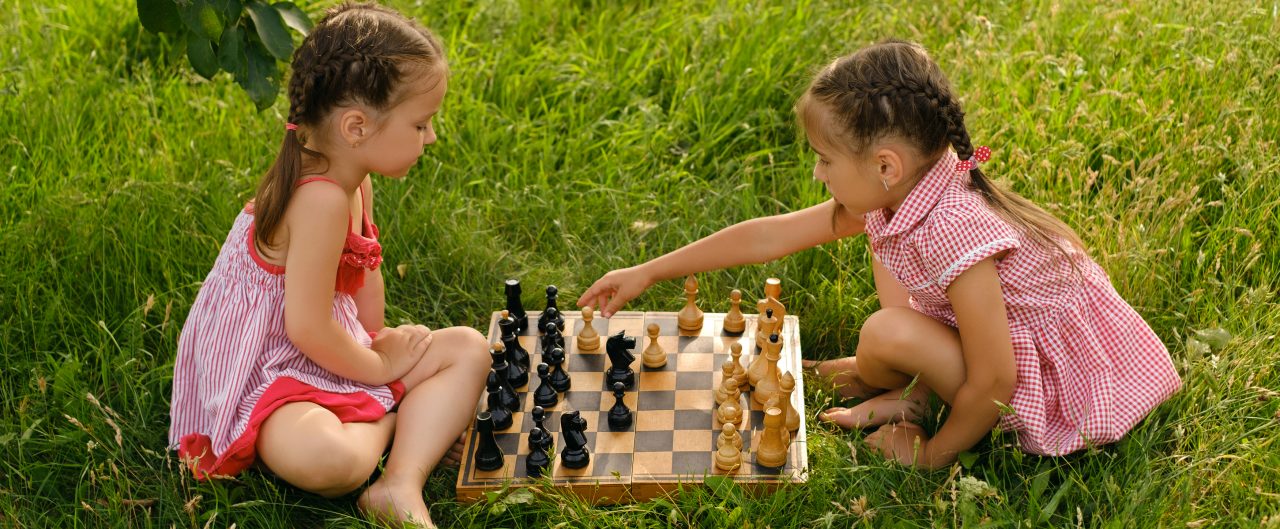 Two girls are playing chess in the garden on the grass. An old wooden chessboard with pieces. 