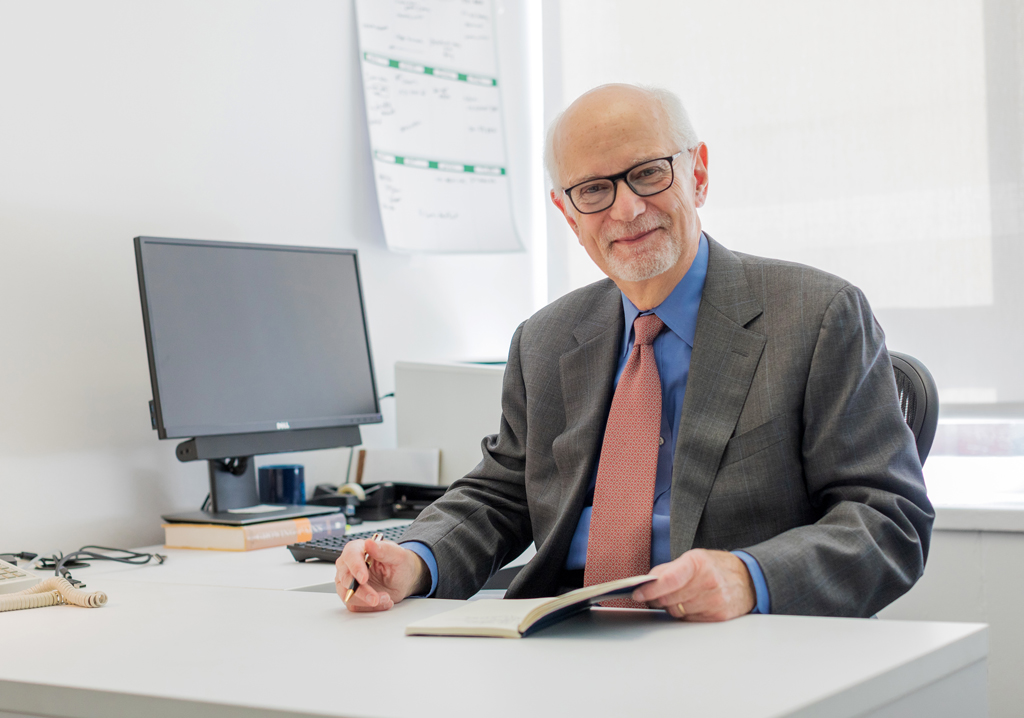 A businessman, with Michael Preston, sitting at his desk in front of computer with pen and paper in his hands