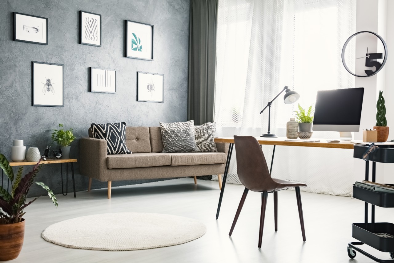 Modern home office with a grey wall with photos hung on the wall, a tan couch with throw pillows, a leather chair, desktop computer on the desk, and black 3-tiered organization system.