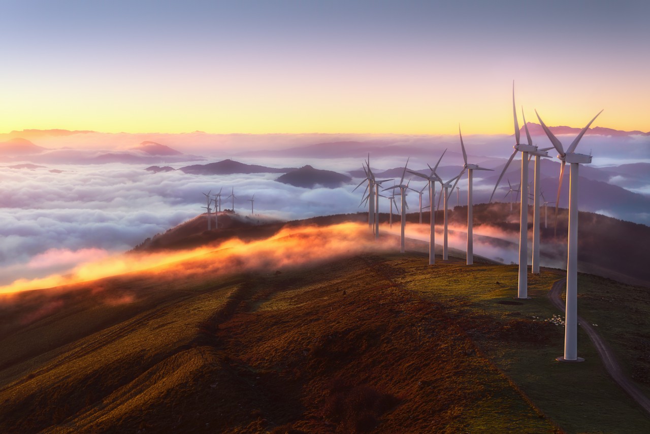 Wind turbines on the top of a mountain seen above clouds and a rising sun.